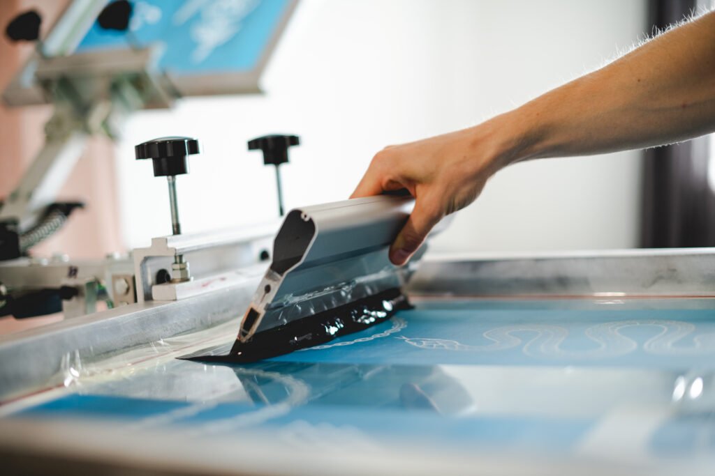 Cropped view of artisan holding squeegee with plastisol ink while working with screen printing machine in designer studio